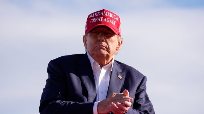 A cleanshaven older man wearing a baseball cap which says Make America Great Again is shown in an  outdoor setting.