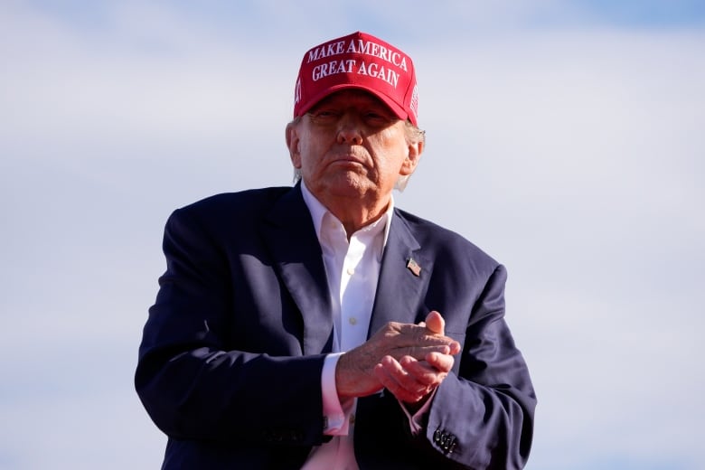 A cleanshaven older man wearing a baseball cap which says Make America Great Again is shown in an  outdoor setting.