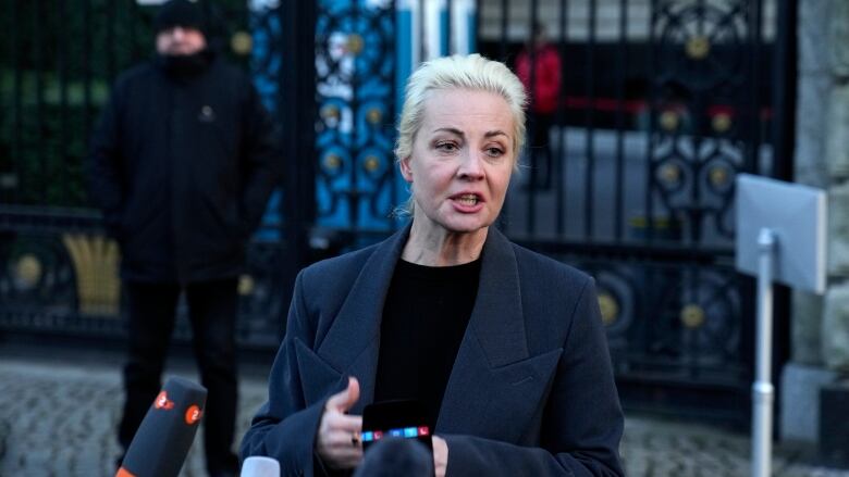 A blonde-haired woman in a blue jacket and black top speaks to reporters on a street.