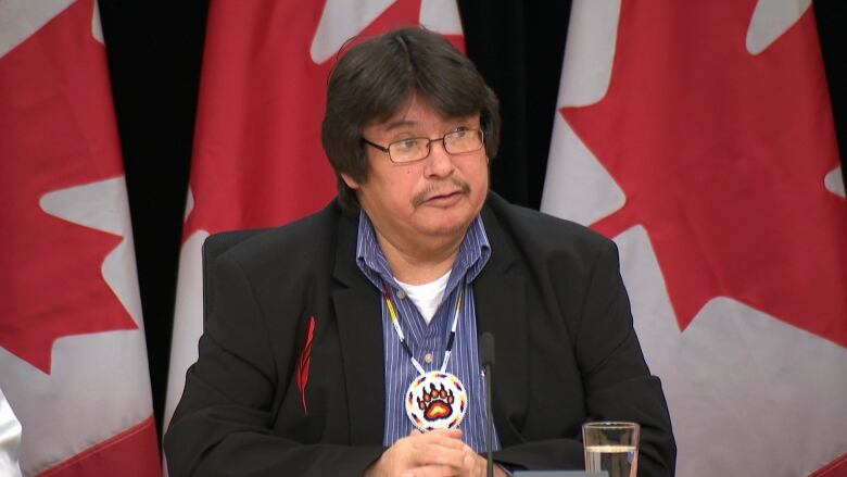 A man in a black suit sitting behind a desk with Canadian flags in the background. 