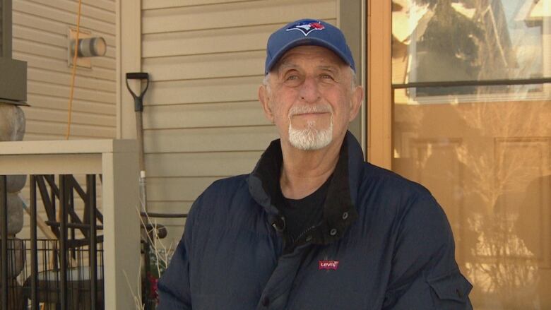 Wayne Bonnar is pictured with his two-storey house in Okotoks, Alta. in the background. He's wearing a blue, down-filled jacket and a Blue Jays baseball cap. 