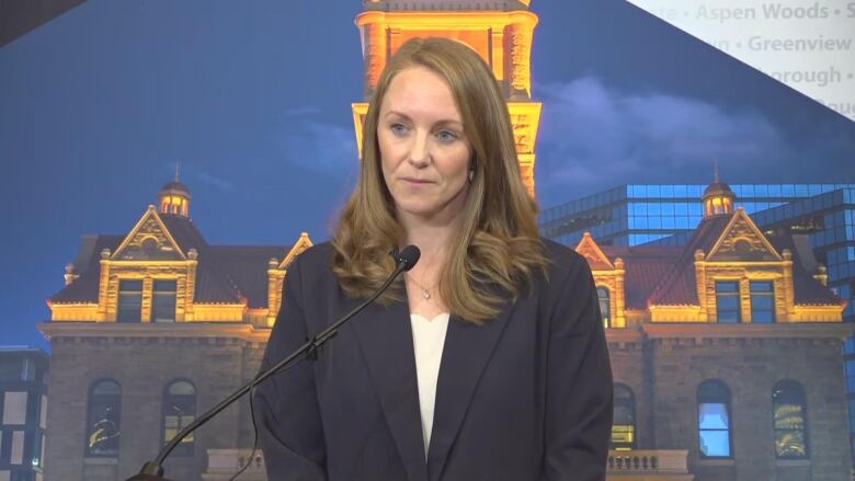 A photo of a woman dressed in a dark blazer at a press conference. She is seen in front of a mic.