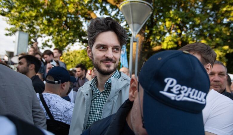 A young man, standing in a crowd, smiles while looking to his left. 