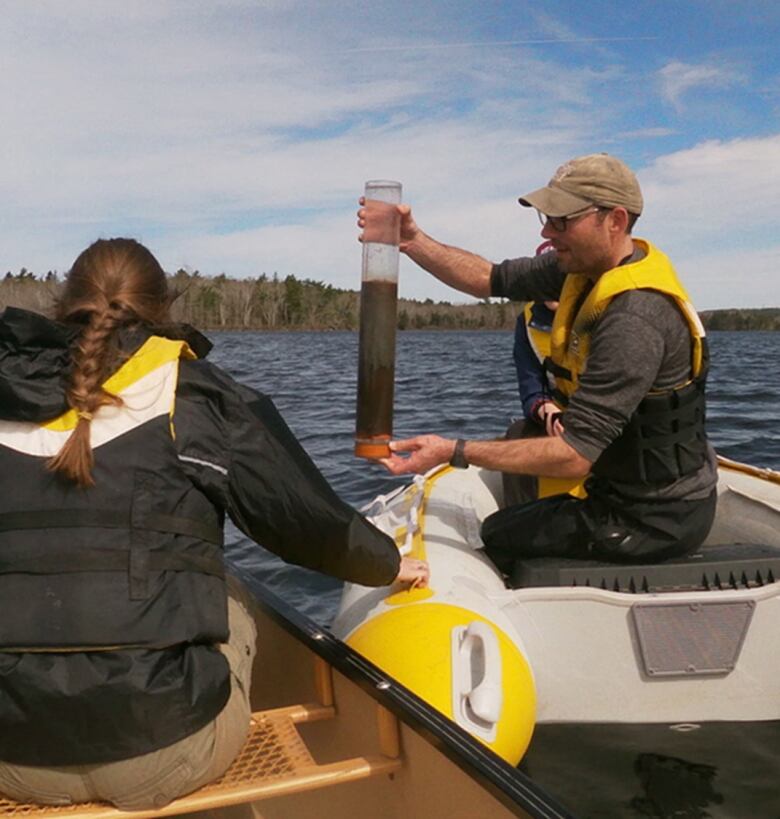 A man wearing a life jacket in a boat holds a large tube, containing a water sample. There is a woman next to him, with one braid, also wearing a life jacket. 