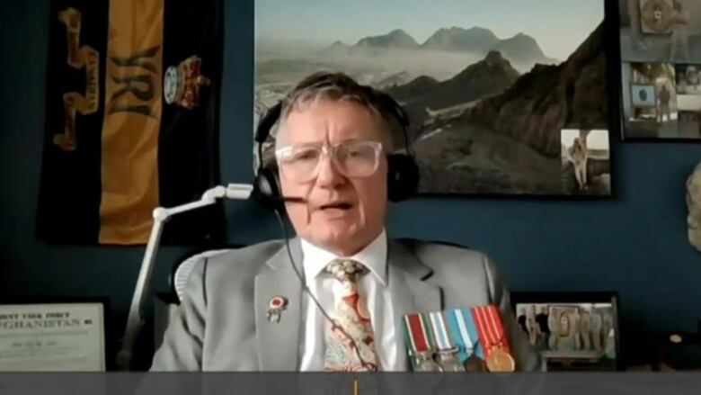 A man with glasses, wearing a grey suit and military medals, speaks into a computer camera.