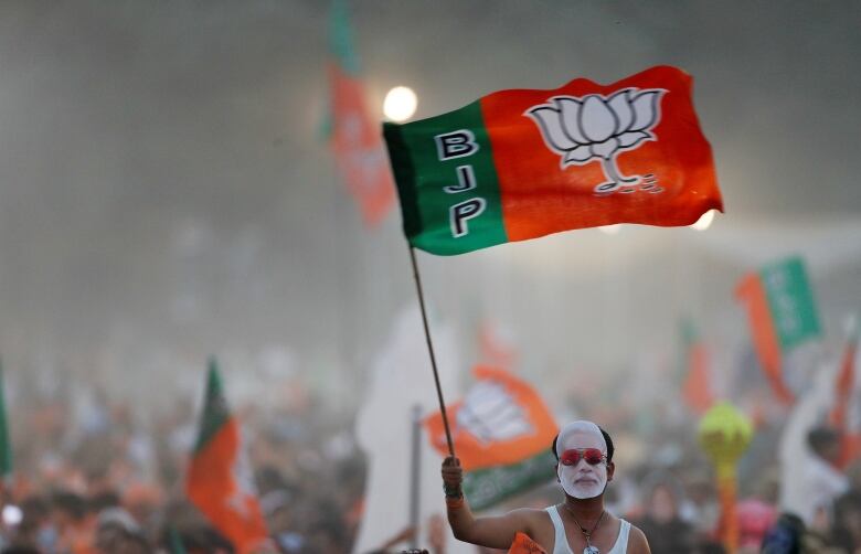 A man wears a paper mask of India's prime minister. He is holding a green and orange flag.