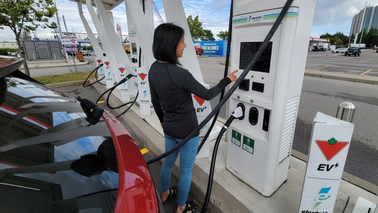 A woman with short dark hair points at the screen of an electric vehicle charging station while her red car is charging. 