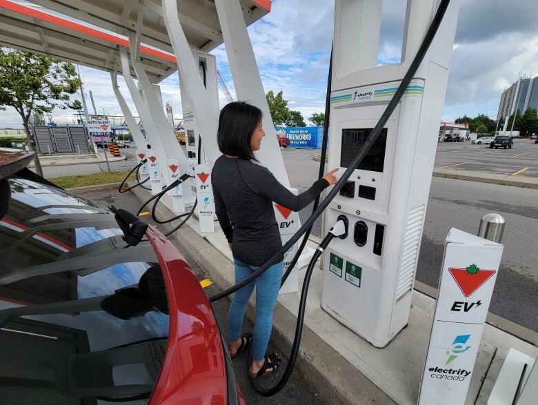 A woman with short dark hair points at the screen of an electric vehicle charging station while her red car is charging. 