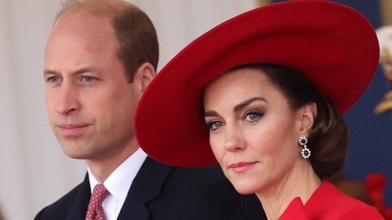 A man in  a suit stands beside a woman in a red hat and blazer, both of them looking serious.