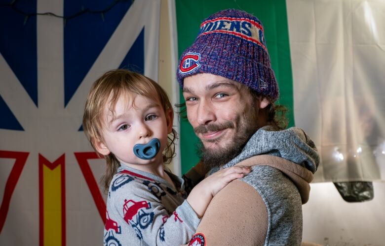 A person wearing a toque holds a young child who has a pacifier in their mouth.