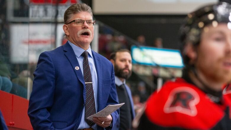 A hockey coach wearing a suit and tie stands on a bench watching his team play 