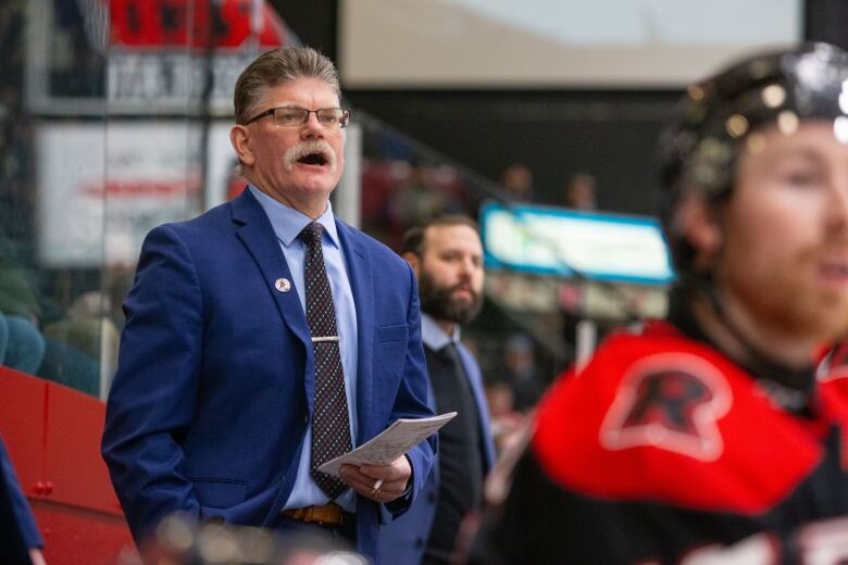 A hockey coach wearing a suit and tie stands on a bench watching his team play 