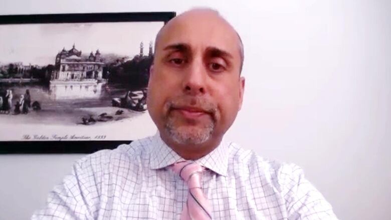 A man wearing a white plaid shirt and a pink tie with stripes on it is seated in front of a white wall with a framed picture behind him. 