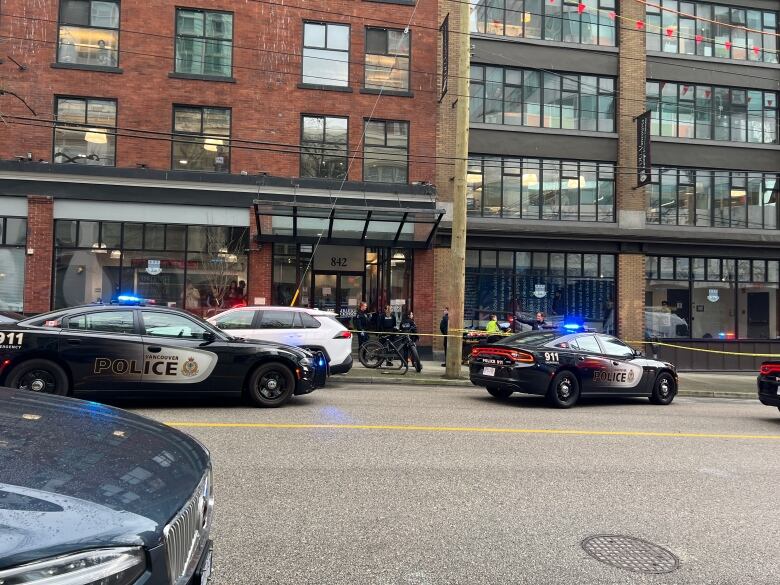 Police cruisers with emergency lights on are parked outside a city business, as officers stand on a sidewalk behind yellow tape.