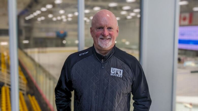 A man with a beard wearing a UPEI hockey shirt stands with the rink in the background