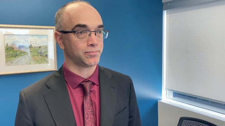 A white man with glasses wearing a grey suit with a red patterned tie and shirt stands in an office with a blue wall behind him