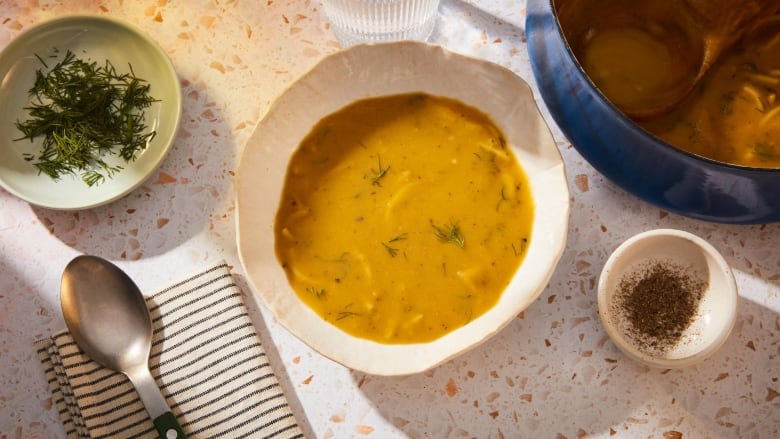 Overhead shot of a white bowl of yellow soup on a terrazzo countertop. A small bowl of dill, a spoon and napkin and a blue Dutch oven surround the bowl. 