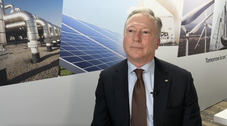 A man sits in front of a large poster showing natural gas pipes, solar panels and wind turbines.