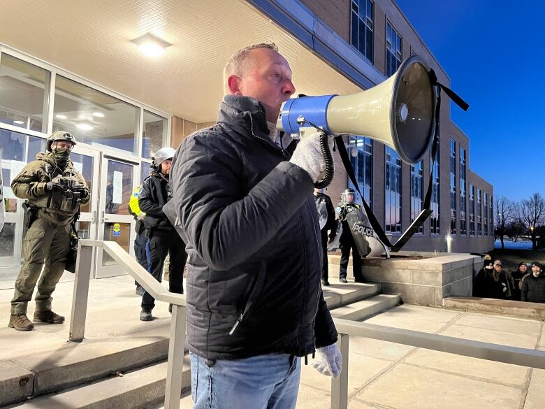 A man with a megaphone standing on stairs with police behind him.