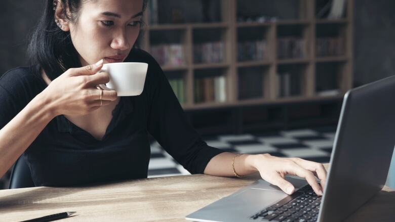 A woman drinks coffee while typing on a laptop in a home office