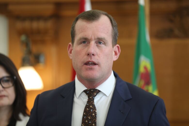 A man in a blue suit and white shirt stands in front of a Saskatchewan flag. 