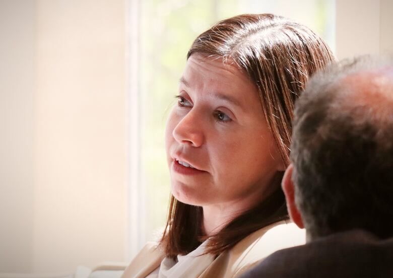 A woman with long brown hair wearing a cream coloured jacket sits in front of a sunny window.