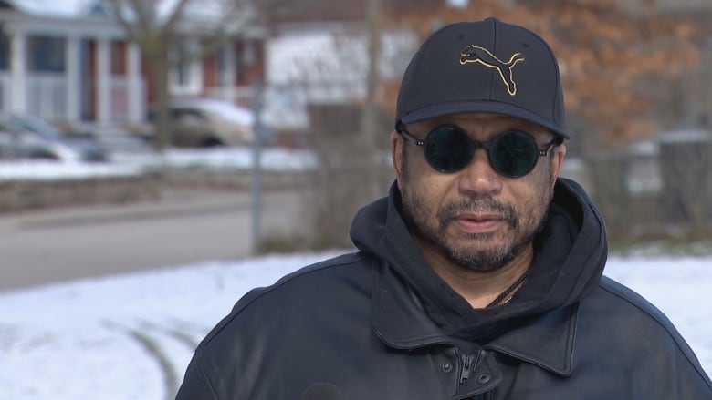 A man wearing all black clothing, a hoodie, jacket and hat, stands in a snowy park.