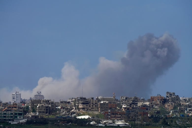 Smoke is shown rising in a clear sky in a distance photo showing a city skyline of low-rise buildings, several which appear to be damaged.