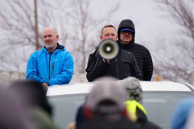 Three men standing in the back of a truck. The guy in the middle is holding a megaphone.