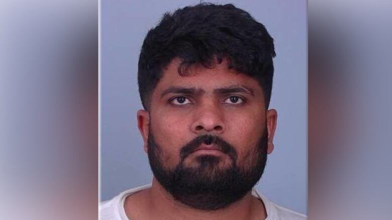 A mugshot of a South Indian man with a black beard wearing a white t-shirt, posing expressionless in front of a plain background.