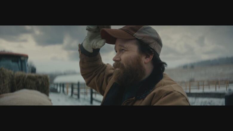 A bearded male actor playing the role of a farmer holds the brim of his cap while looking off into the distance. 