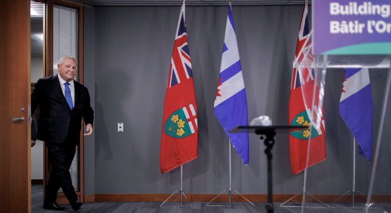 Premier Doug Ford steps through a doorway into a room with Ontario and Toronto flags in the background.  