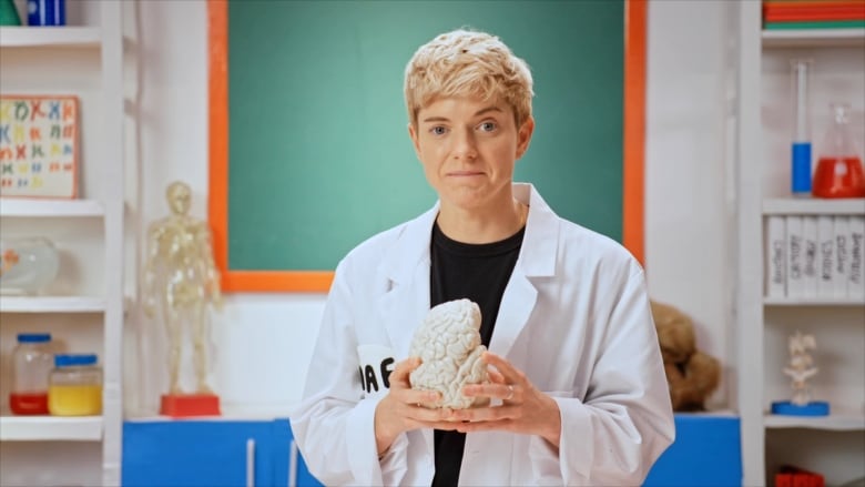 A person with short blond hair, wearing a lab coat, holds a white model of a human brain.