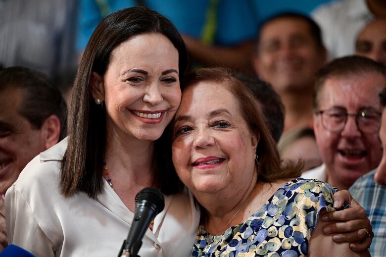 Two people embrace in front of a microphone as people gather behind.