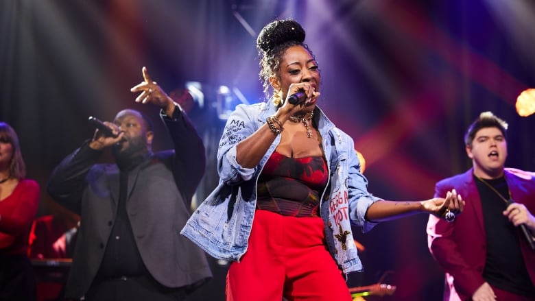 Jah'mila (a Black woman wearing a denim jacket and red pants) stands in centre stage, singing with a microphone in her hand. Owen O'Sound Lee (a Black man) is to her left and Wolf Castle (an Indigneous man) is to her right. 