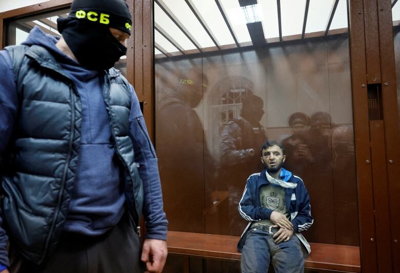 A person with facial injuries sits behind a glass wall as a person wearing a balaclava watched them.