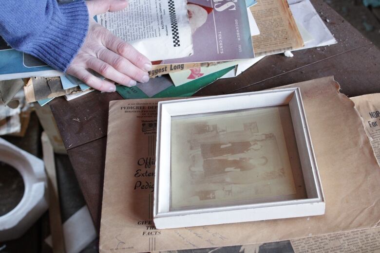 A picture frame with an old photograph sits on a table with a woman's hand next to it on a pile of papers.