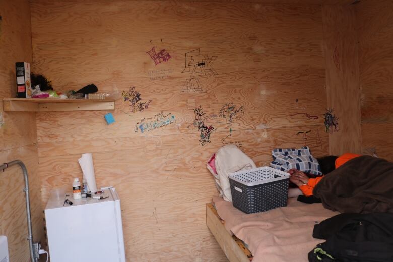 A woman lies in bed in a tiny bare room with grafitti on the walls. 