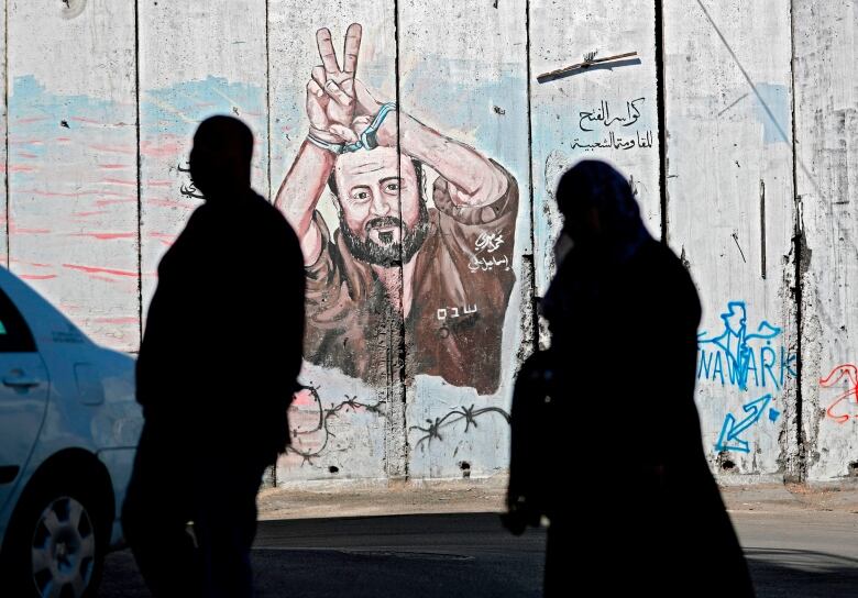 Two people in silhouette walk past a wall with graffiti and a portrait of a man.