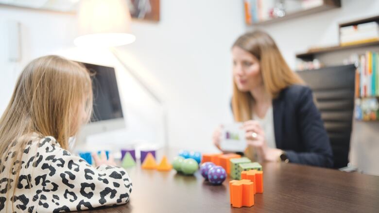 A child with long hair leans on a desk with a row of wooden blocks set in front of them. A woman on the opposite side holds a card. 