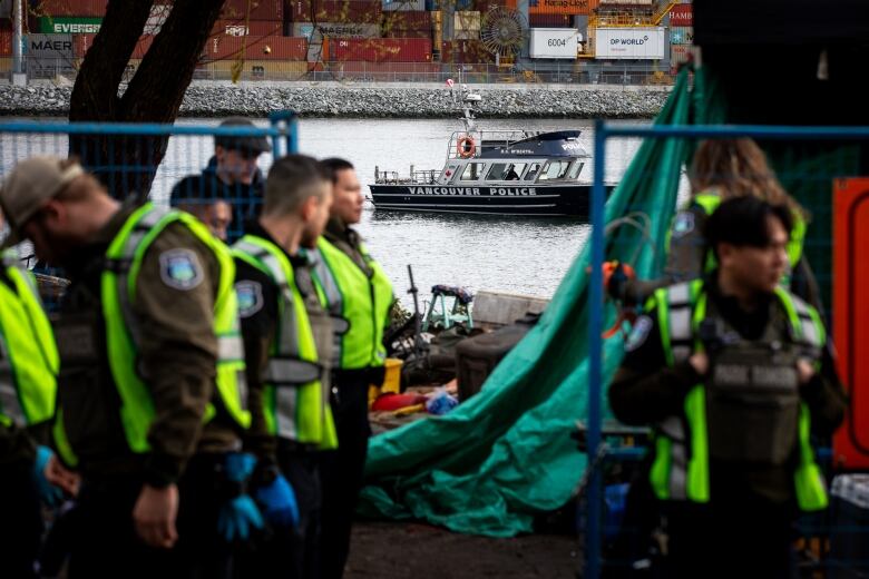 Park rangers wearing high-visibility vests pose at a homeless encampment, with a police boat visible in the background.