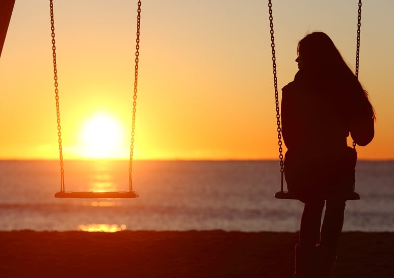 A silhouette of a woman on a swing, facing a setting sun. The other swing is empty. 