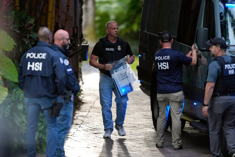 Five men wearing jackets, shirts and bulletproof vests with the words 'Police HSI' stand around a van. The man in the middle holds a large plastic bag with a rectangular object inside.