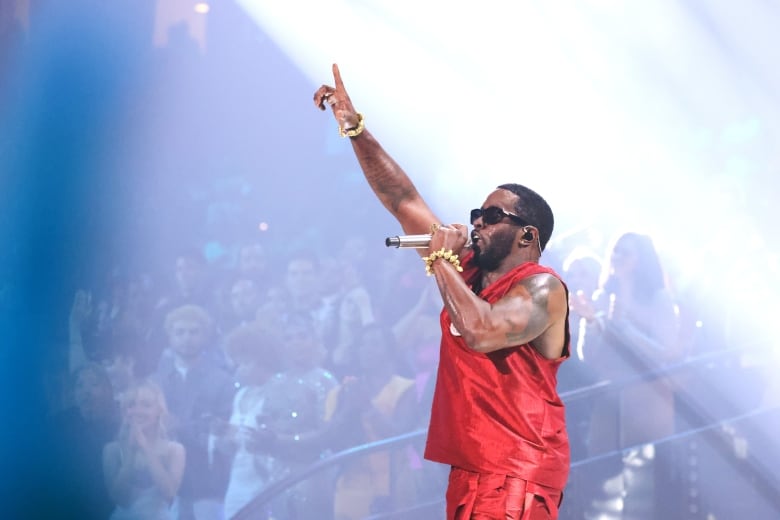 A man wearing a red shirt and pants holds his hand up while singing into a microphone onstage.