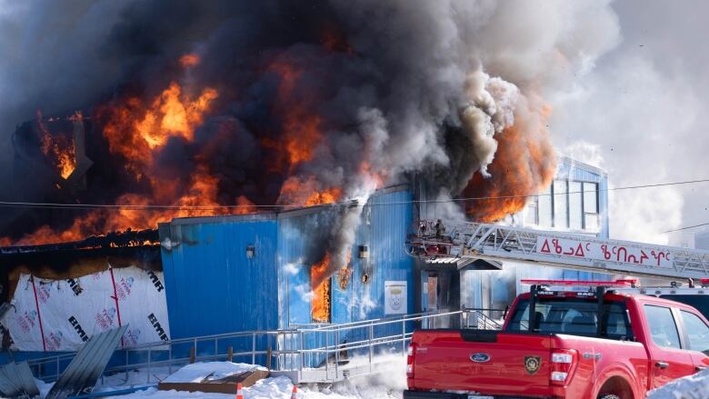 Flames and smoke come out of a blue building with a fire truck in front of it 