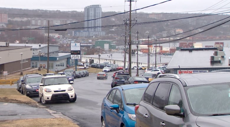 Cars are lined along both sides of the road with low-rise commercial buildings along the hill, with a view of the water in the distance