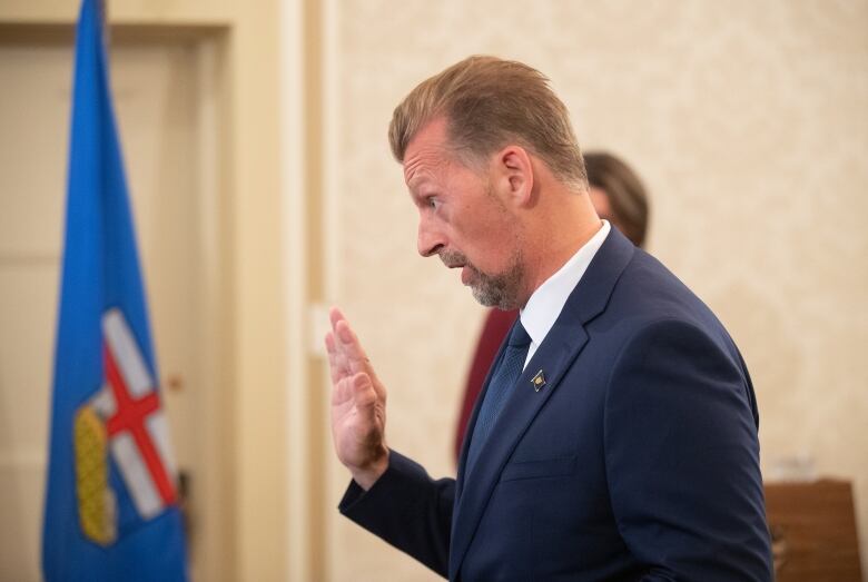 A man with blonde hair and a goatee in a blue suit raises his right hand.