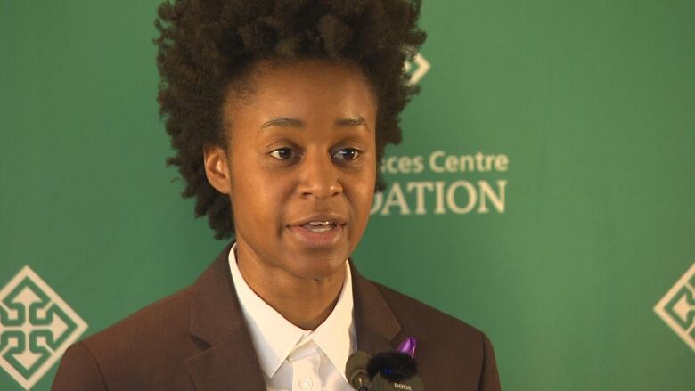 A person in a brown suit and white shirt stand behind a microphone and in front of a green backdrop. 