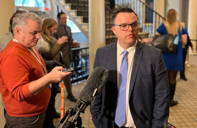 A man with glasses in a suit and tie stands at a microphone as people stand around him.