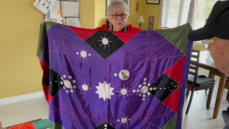 A woman with short, white hair holds up a giant purple flag patterned with suns and moons.